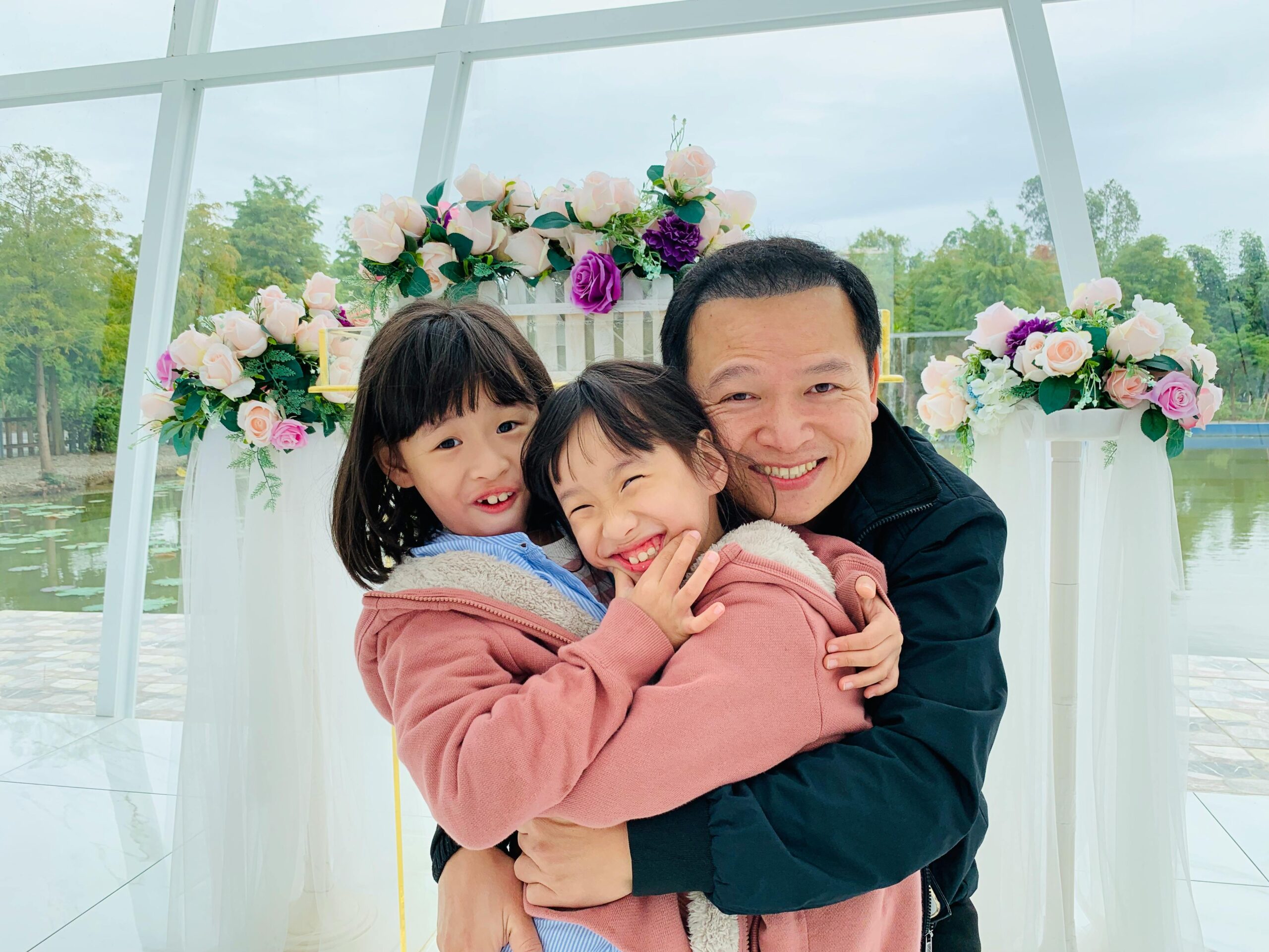 pexels-photo-3887653-3887653 Father and daughters sharing joyful embraces and smiles in a floral setting.
