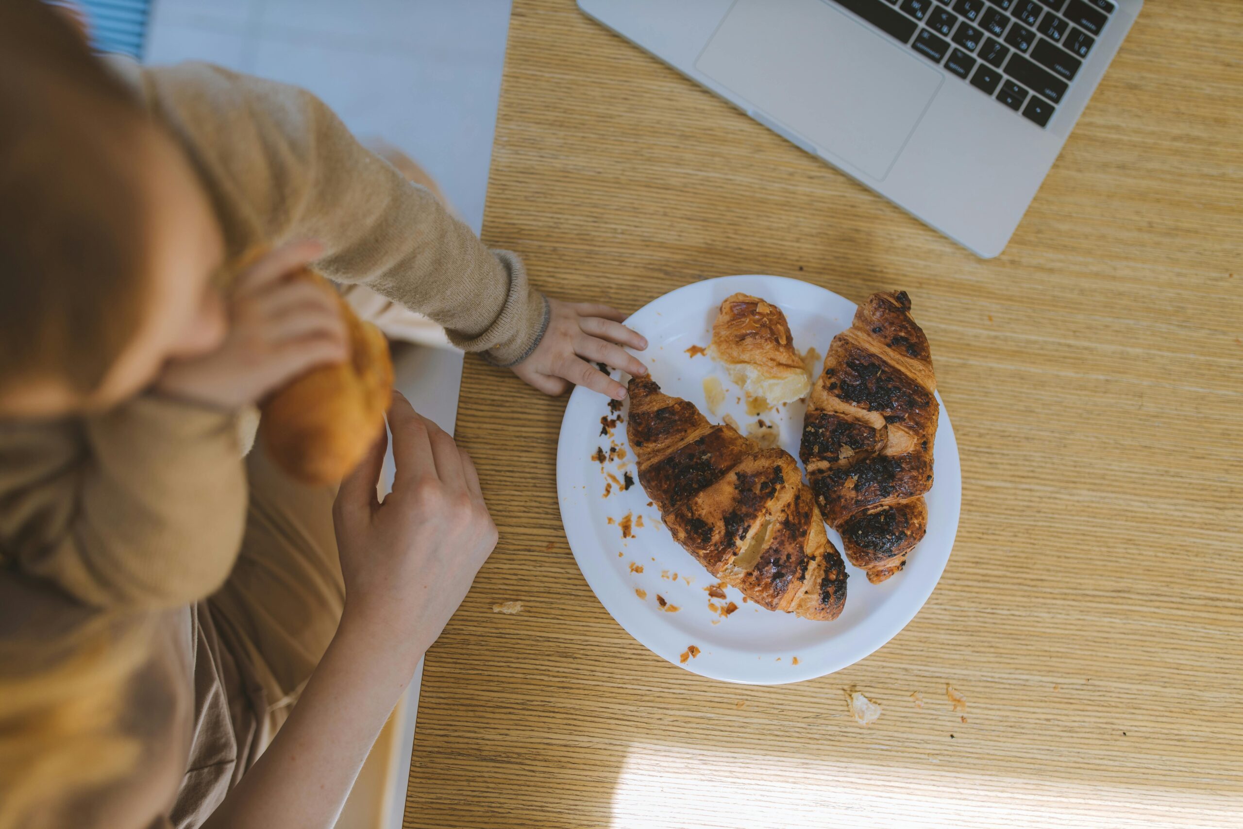 Overhead view of a child enjoying croissants at a table with a laptop nearby, capturing a cozy breakfast moment.