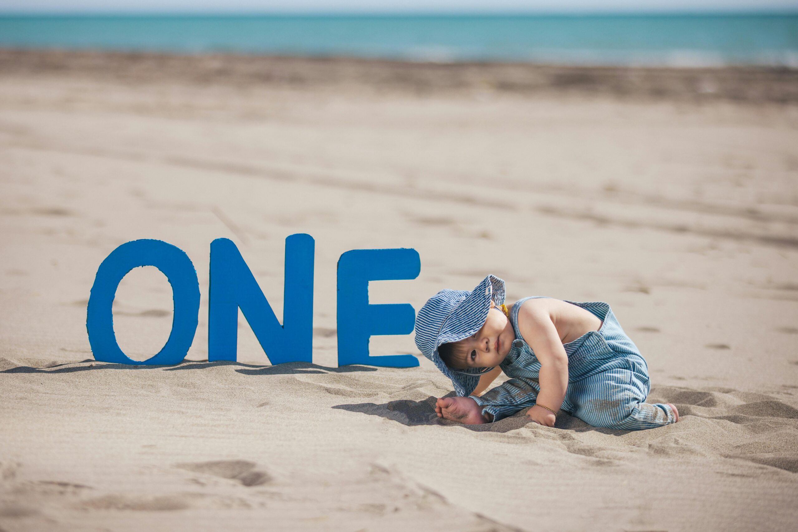 Toddler celebrating first birthday at sandy beach with 'ONE' sign, wearing a hat.